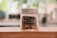 A glass jar with a silver lid contains reddish-brown chilli flakes. The jar is labeled with black tape displaying the words 'CHILLI FLAKES.' It is placed on a wooden surface, with a blurred, green and brown background suggesting a natural or kitchen setting.