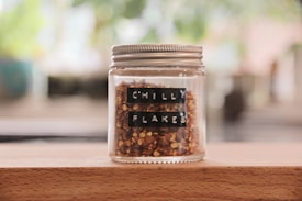 A glass jar with a silver lid contains reddish-brown chilli flakes. The jar is labeled with black tape displaying the words 'CHILLI FLAKES.' It is placed on a wooden surface, with a blurred, green and brown background suggesting a natural or kitchen setting.
