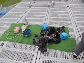 Climbing gear including helmets, harnesses, ropes, and other equipment is arranged on a green mat placed over a metal grid surface. The scene includes climbing helmets in blue and various metal carabiners and tools. A person's leg is visible on the right side, suggesting preparation or presence of a person for an upcoming activity.