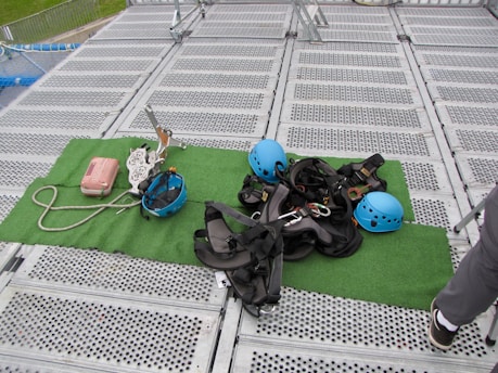 Climbing gear including helmets, harnesses, ropes, and other equipment is arranged on a green mat placed over a metal grid surface. The scene includes climbing helmets in blue and various metal carabiners and tools. A person's leg is visible on the right side, suggesting preparation or presence of a person for an upcoming activity.