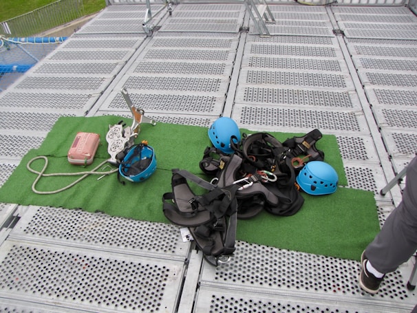 Climbing gear including helmets, harnesses, ropes, and other equipment is arranged on a green mat placed over a metal grid surface. The scene includes climbing helmets in blue and various metal carabiners and tools. A person's leg is visible on the right side, suggesting preparation or presence of a person for an upcoming activity.