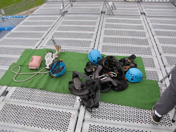 Climbing gear including helmets, harnesses, ropes, and other equipment is arranged on a green mat placed over a metal grid surface. The scene includes climbing helmets in blue and various metal carabiners and tools. A person's leg is visible on the right side, suggesting preparation or presence of a person for an upcoming activity.