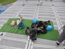 Climbing gear including helmets, harnesses, ropes, and other equipment is arranged on a green mat placed over a metal grid surface. The scene includes climbing helmets in blue and various metal carabiners and tools. A person's leg is visible on the right side, suggesting preparation or presence of a person for an upcoming activity.