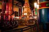 An indoor amusement park scene with a vibrant ride structure resembling a miniature Eiffel Tower. The area is adorned with colorful columns and metallic fixtures, while amusement ride seats are visible on the left. The environment features a variety of light sources contributing to a festive atmosphere.