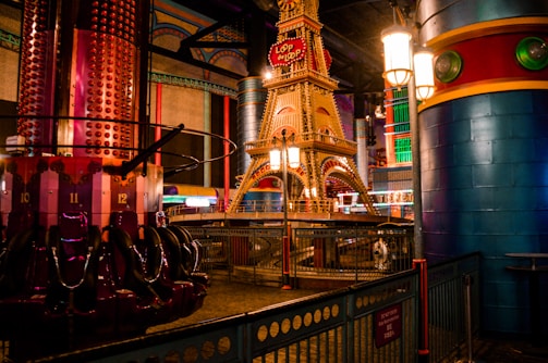 An indoor amusement park scene with a vibrant ride structure resembling a miniature Eiffel Tower. The area is adorned with colorful columns and metallic fixtures, while amusement ride seats are visible on the left. The environment features a variety of light sources contributing to a festive atmosphere.