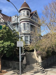 A multi-story Victorian-style house with intricate architectural details, featuring a prominent turret and ornate trim. A set of stone steps leads up to the entrance, surrounded by lush greenery and a bare tree. Street signs are visible, indicating a speed limit and a restriction on tour buses.