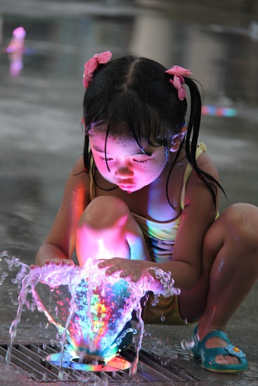 A child with wet hair and pink bows crouches by a fountain, intensely focused on the colorful, illuminated water spraying upwards. The child is wearing a yellow swimsuit and blue sandals, and the surrounding area is wet, indicating a playful and refreshing environment.