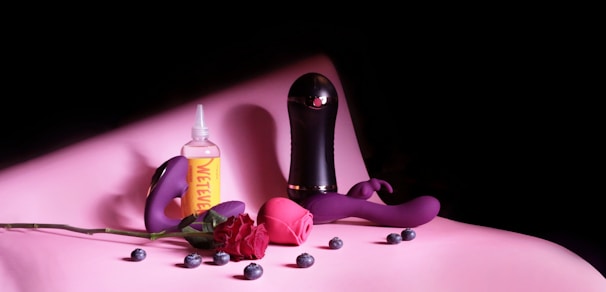 A collection of items is elegantly arranged on a soft pink surface. There is a bottle of lubricant labeled 'WET' alongside two vibrators, with blueberries scattered around them. A red rose lies across the scene, adding a romantic touch.
