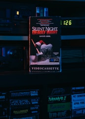 A VHS tape titled 'Silent Night, Deadly Night' with a Santa figure holding an axe on the cover. The scene is dimly lit with a blue and red hue, set on a shelf with other VHS tapes like 'Halloween Resurrection' and 'Pumpkinhead II'. An old electronic device with a digital clock displaying 1:26 is visible in the background.