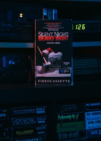 A VHS tape titled 'Silent Night, Deadly Night' with a Santa figure holding an axe on the cover. The scene is dimly lit with a blue and red hue, set on a shelf with other VHS tapes like 'Halloween Resurrection' and 'Pumpkinhead II'. An old electronic device with a digital clock displaying 1:26 is visible in the background.