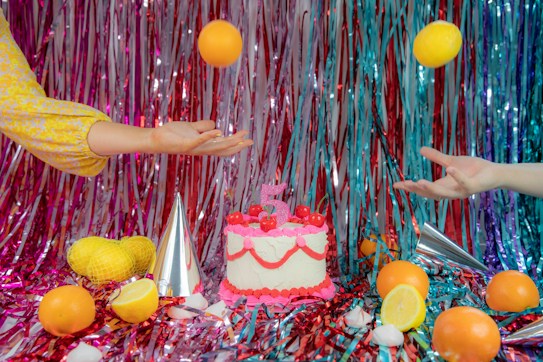 Two hands, one on each side, are playfully tossing an orange and a lemon in the air. In the center, a decorated cake with layers of pink frosting and cherries sits on a festive surface covered with shiny, colorful tinsel strips in shades of red, pink, and blue. Several citrus fruits and metallic party hats are scattered around.