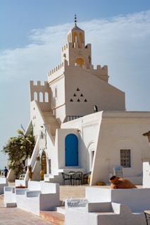 A white-washed Mediterranean-style building with blue doors and windows, featuring distinct architectural elements like arches and triangular cutouts. The setting includes chairs and tables outside, with potted plants and a large amphora. The sky is partly cloudy, adding a serene backdrop.