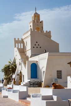 A white-washed Mediterranean-style building with blue doors and windows, featuring distinct architectural elements like arches and triangular cutouts. The setting includes chairs and tables outside, with potted plants and a large amphora. The sky is partly cloudy, adding a serene backdrop.