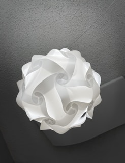 A white, geometric, origami-style lampshade hanging from the ceiling. The lampshade features intricate curves and swirls, creating a modern and artistic appearance. The ceiling is textured and gray, providing a neutral background that contrasts with the bright lamp.
