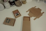 A white desk features various crafting items, including an assembled cardboard box with mountain-themed decorations, an open box displaying a pair of decorative earrings with red flowers, and scattered flat cardboard pieces intended for box-making. Surrounding these items are jars and containers holding string, dried flowers, tools, and other crafting supplies.