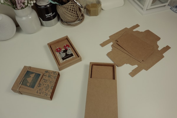 A white desk features various crafting items, including an assembled cardboard box with mountain-themed decorations, an open box displaying a pair of decorative earrings with red flowers, and scattered flat cardboard pieces intended for box-making. Surrounding these items are jars and containers holding string, dried flowers, tools, and other crafting supplies.