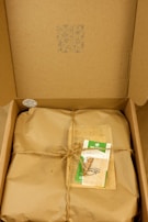 A cardboard box containing a wrapped package tied with twine. On top of the package, there is a card with green accents and a design featuring an eye symbol. The box's lid has patterns and symbols printed in blue ink.
