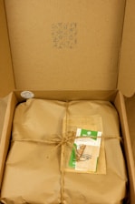 A cardboard box containing a wrapped package tied with twine. On top of the package, there is a card with green accents and a design featuring an eye symbol. The box's lid has patterns and symbols printed in blue ink.