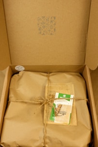 A cardboard box containing a wrapped package tied with twine. On top of the package, there is a card with green accents and a design featuring an eye symbol. The box's lid has patterns and symbols printed in blue ink.