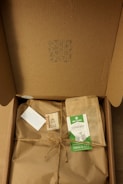 A cardboard box opened to reveal brown paper wrapping tied with twine. There is an eco-friendly label, a small card, and an envelope on top of the wrapping. The inside lid has a small pattern printed on it.