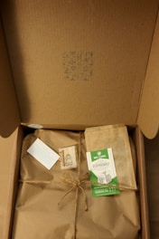 A cardboard box opened to reveal brown paper wrapping tied with twine. There is an eco-friendly label, a small card, and an envelope on top of the wrapping. The inside lid has a small pattern printed on it.