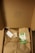 A cardboard box opened to reveal brown paper wrapping tied with twine. There is an eco-friendly label, a small card, and an envelope on top of the wrapping. The inside lid has a small pattern printed on it.