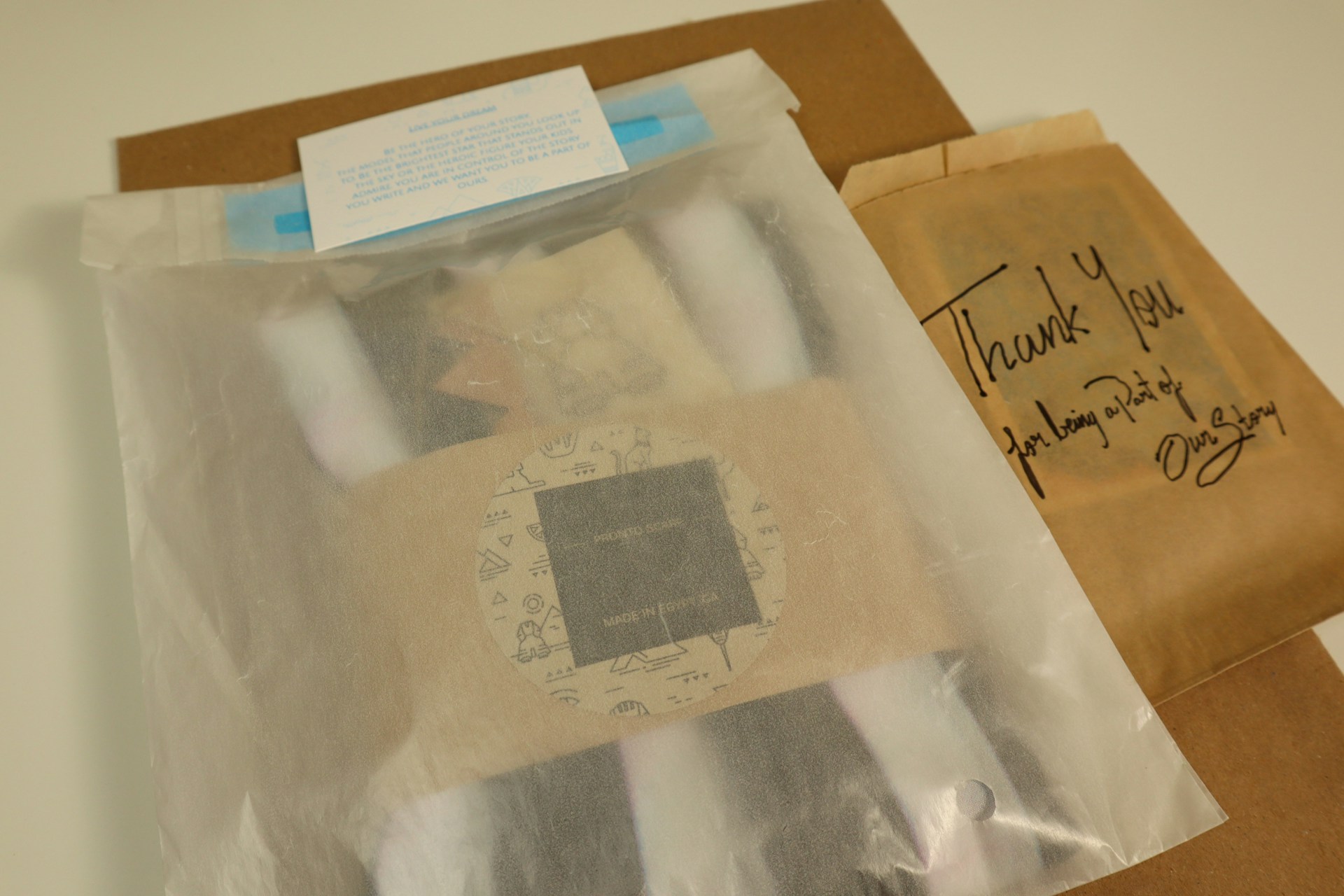 A collection of items is wrapped in semi-transparent packaging, with a visible circular label featuring various illustrations. Beside it, a brown paper bag has 'Thank You' written on it in black marker, accompanied by a handwritten note expressing gratitude for being part of a story.