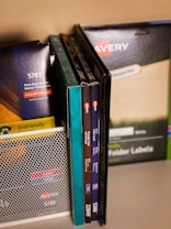 A collection of office supplies neatly arranged, including Avery label and card packages. The items are stored on a shelf and organized upright against a mesh divider. Various colors and sizes of packaging are visible, with text such as 'EcoFriendly' and product numbers.