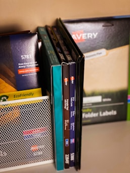 A collection of office supplies neatly arranged, including Avery label and card packages. The items are stored on a shelf and organized upright against a mesh divider. Various colors and sizes of packaging are visible, with text such as 'EcoFriendly' and product numbers.