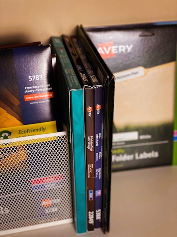 A collection of office supplies neatly arranged, including Avery label and card packages. The items are stored on a shelf and organized upright against a mesh divider. Various colors and sizes of packaging are visible, with text such as 'EcoFriendly' and product numbers.