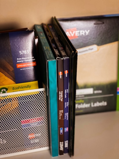 A collection of office supplies neatly arranged, including Avery label and card packages. The items are stored on a shelf and organized upright against a mesh divider. Various colors and sizes of packaging are visible, with text such as 'EcoFriendly' and product numbers.