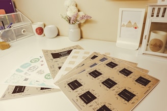 A neatly arranged desk with several stationary items including sheets of paper with various designs and labels. A small bouquet of flowers sits in a white vase, and there's a framed picture of geometric mountains. A white speaker and a white cup with a red pattern are positioned near a wire mesh organizer. A decorative lantern and a mug with a mountain design are also present.