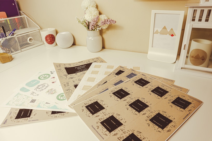 A neatly arranged desk with several stationary items including sheets of paper with various designs and labels. A small bouquet of flowers sits in a white vase, and there's a framed picture of geometric mountains. A white speaker and a white cup with a red pattern are positioned near a wire mesh organizer. A decorative lantern and a mug with a mountain design are also present.