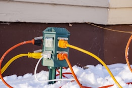 A green outdoor electrical outlet is surrounded by various brightly colored extension cords plugged into it. The outlet and cords are set against a brown wall, with snow covering the ground around them.