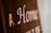 Blurry close-up of text on a brown surface with white lettering. The text reads 'A Home' with additional, partially obscured words.