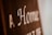 Blurry close-up of text on a brown surface with white lettering. The text reads 'A Home' with additional, partially obscured words.