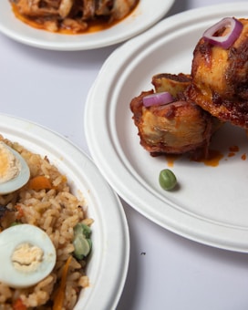Plates of food featuring fried rice mixed with vegetables and garnished with sliced boiled eggs, alongside a serving of grilled chicken topped with red onion slices. Another dish appears in the background, possibly containing a saucy, meaty dish.