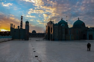 A beautiful mosque with intricate architecture is set against a dramatic sunset sky. The courtyard is spacious and tiled, with a few people visible in the distance. The sky is filled with scattered clouds illuminated by the golden and orange hues of the setting sun.
