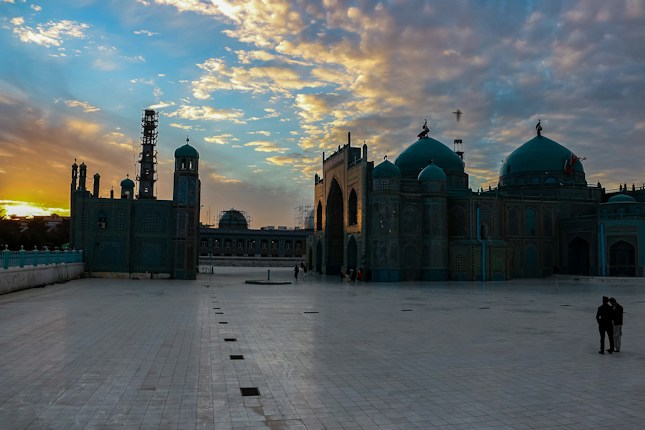 A beautiful mosque with intricate architecture is set against a dramatic sunset sky. The courtyard is spacious and tiled, with a few people visible in the distance. The sky is filled with scattered clouds illuminated by the golden and orange hues of the setting sun.