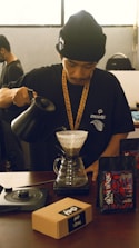 A person wearing a black beanie and black t-shirt is preparing pour-over coffee using a dripper and a kettle. The person is focused on pouring the hot water over the coffee grounds. On the table, there are two bags of coffee beans and a brown box labeled 'Kopi' alongside a coffee scale.