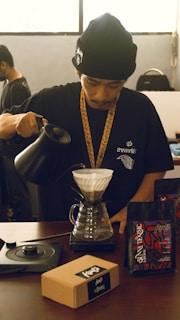 A person wearing a black beanie and black t-shirt is preparing pour-over coffee using a dripper and a kettle. The person is focused on pouring the hot water over the coffee grounds. On the table, there are two bags of coffee beans and a brown box labeled 'Kopi' alongside a coffee scale.
