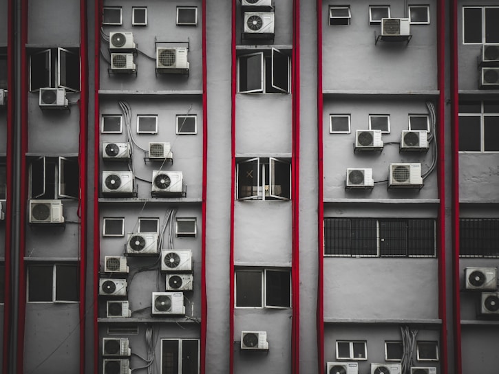 Multiple air conditioning units are mounted on the exterior of a tall building's facade. The structure features numerous small rectangular windows, many of which are open. Vertical red lines divide the facade, creating a repetitive pattern. The overall look is industrial and urban.