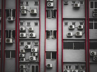 Multiple air conditioning units are mounted on the exterior of a tall building's facade. The structure features numerous small rectangular windows, many of which are open. Vertical red lines divide the facade, creating a repetitive pattern. The overall look is industrial and urban.