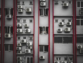 Multiple air conditioning units are mounted on the exterior of a tall building's facade. The structure features numerous small rectangular windows, many of which are open. Vertical red lines divide the facade, creating a repetitive pattern. The overall look is industrial and urban.
