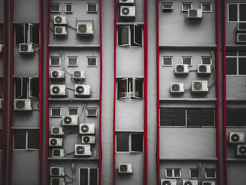 Multiple air conditioning units are mounted on the exterior of a tall building's facade. The structure features numerous small rectangular windows, many of which are open. Vertical red lines divide the facade, creating a repetitive pattern. The overall look is industrial and urban.