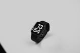 A black smartwatch with a sleek design features a large digital display showing the time as 13:54. It is positioned on a smooth, light gray surface, with the watch and its band elegantly curving in a minimalist setting.