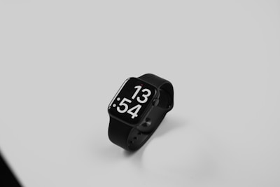 A black smartwatch with a sleek design features a large digital display showing the time as 13:54. It is positioned on a smooth, light gray surface, with the watch and its band elegantly curving in a minimalist setting.