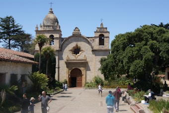 A historic stone church with two bell towers and a detailed facade sits under a clear blue sky. Palm trees and lush greenery surround the building, providing a vibrant contrast. Several people are walking and standing in front of the church, enjoying the day.