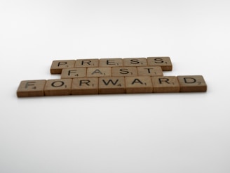 Scrabble tiles are arranged on a white surface to spell out the phrase 'PRESS FAST FORWARD'. The tiles are made of wood and display black letters and numbers.