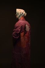 A person is shown in profile, wearing a headscarf with a floral pattern and a long purple coat featuring an artistic depiction of a woman's face and floral designs on the back. The overall lighting is dark and moody, emphasizing the colors and patterns on the clothing.